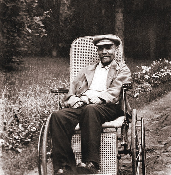An elderly man in a wheel chair, wearing a hat and suit, sits in a garden with flowers and trees in the background.