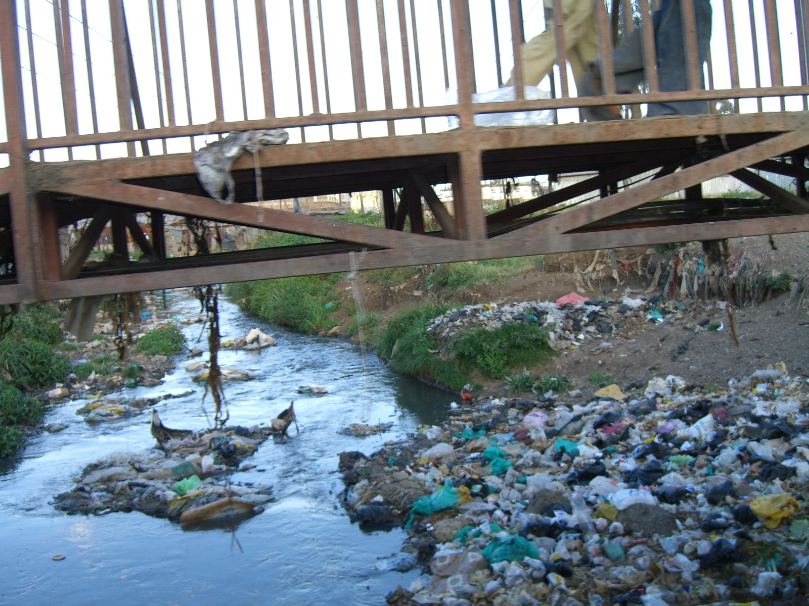 A wooden bridge spans a polluted stream heavily littered with garbage. The water is surrounded by rubbish and debris on its banks.