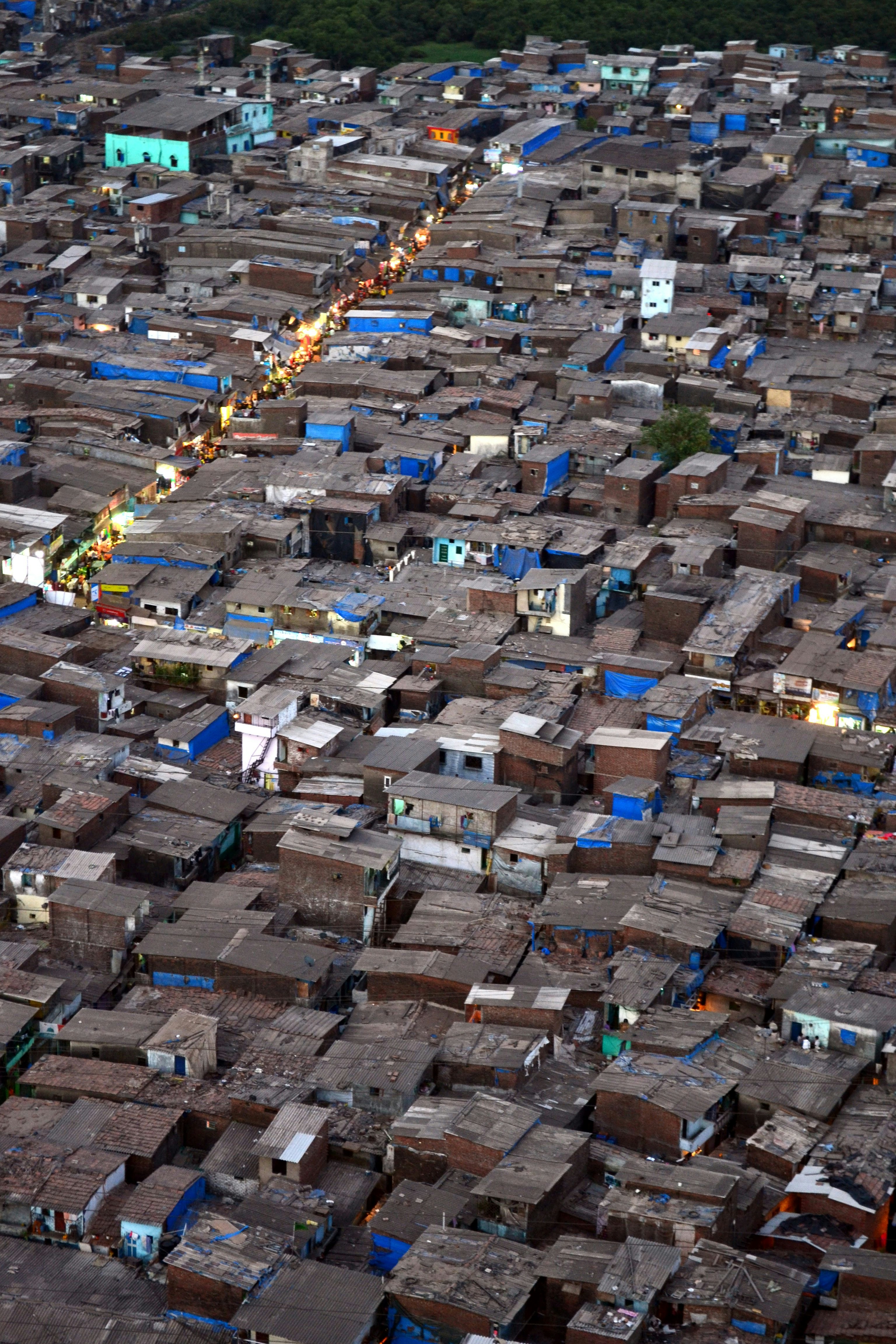 Aerial view of densely packed informal housing with narrow pathways, blue tarps, and bright lights showing a trading street within the settlement.
