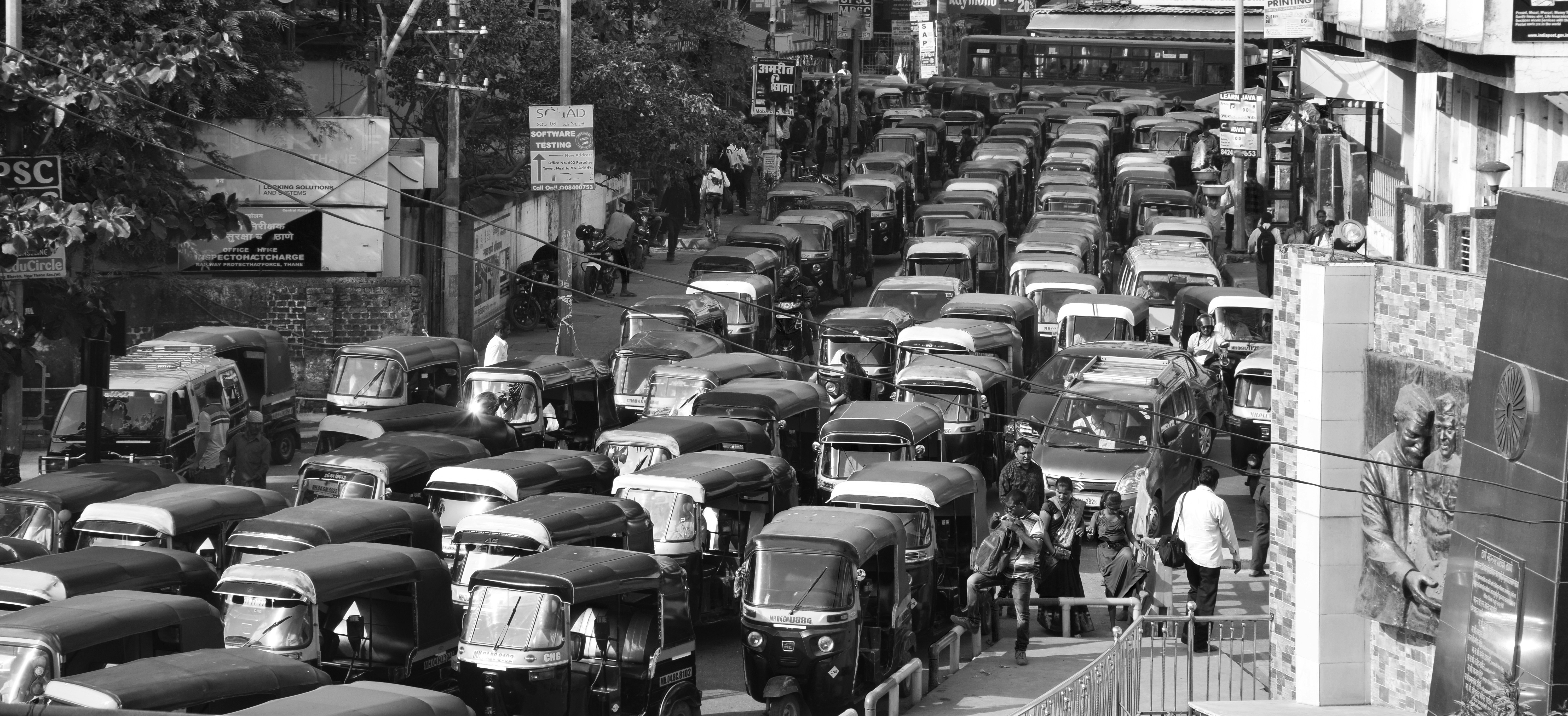 Heavy traffic congestion with numerous auto rickshaws (tuk-tuks) on a busy urban road in Mumbai. People walk along the pavement. Trees and buildings line the road.
