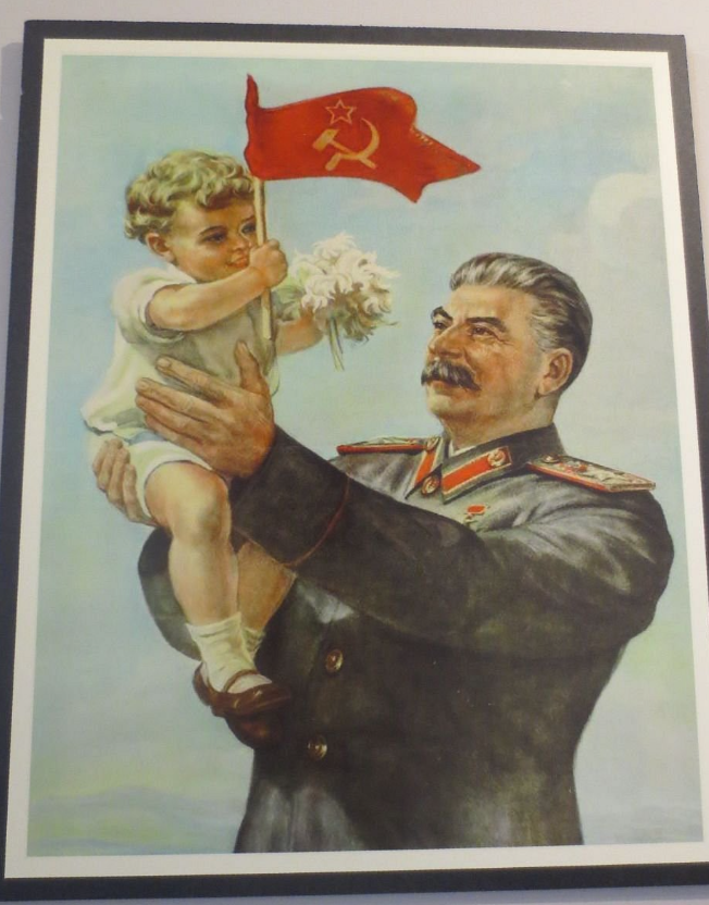 A man in a military uniform (Stalin) holds a child with flowers and a small red flag featuring a hammer and sickle against a light backdrop.