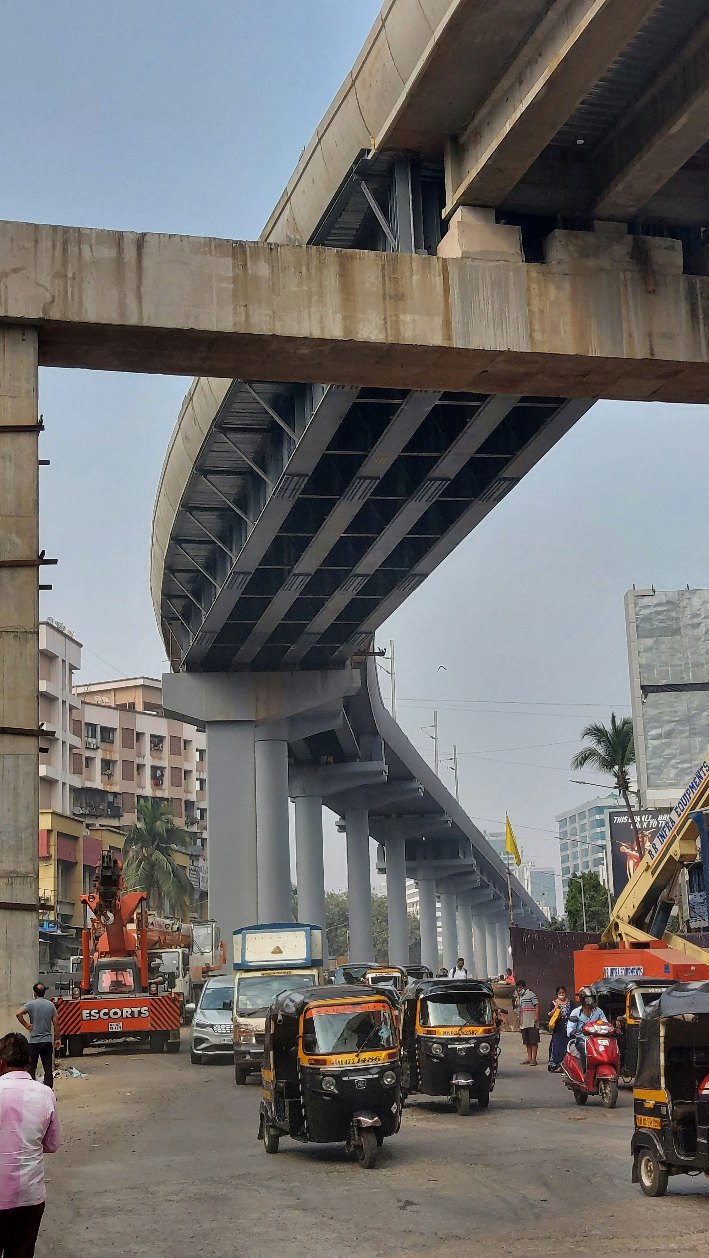 Busy street with auto rickshaws under a concrete metro flyover under construction. Buildings and palm trees are visible in the background.