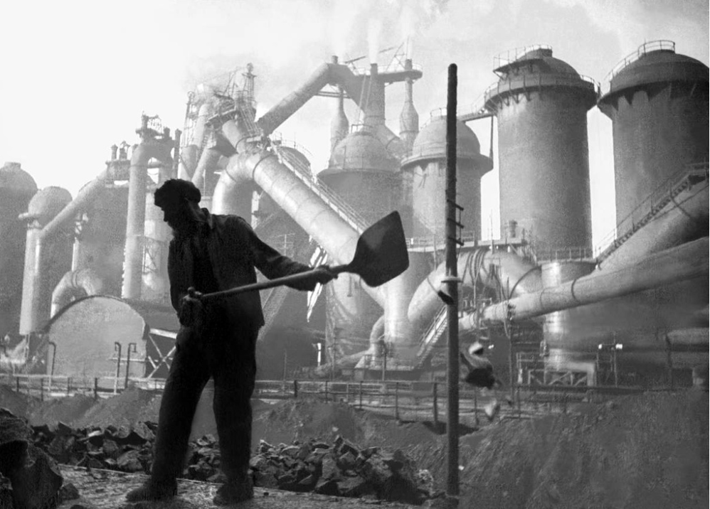 Silhouetted worker with a shovel in front of large industrial chimneys and machinery, emitting smoke, suggesting an active factory environment.