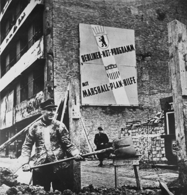 Worker digging at a construction site in post-war Berlin, wearing a cap and coat. Background shows a banner promoting the Marshall Plan aid.